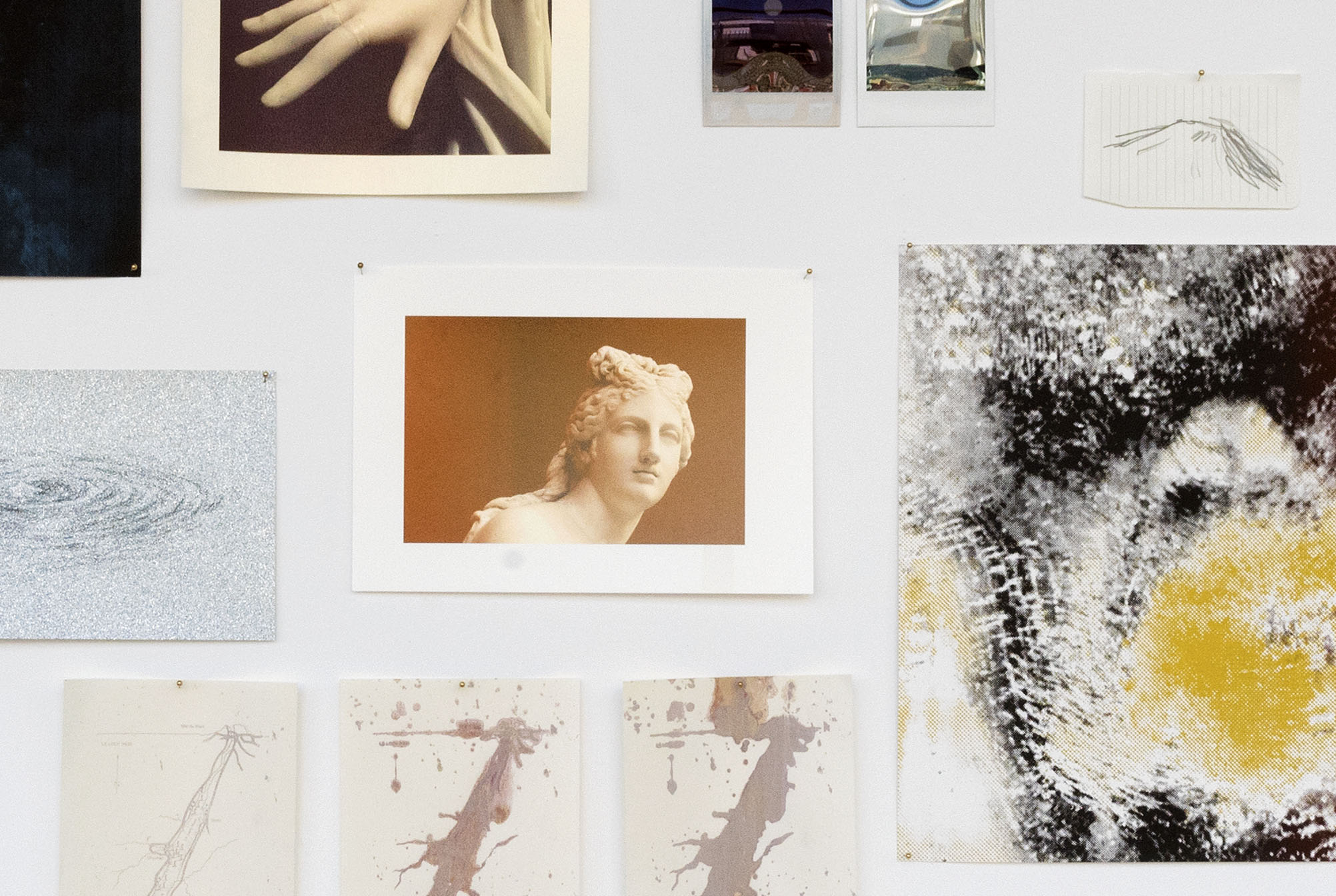 Gros plan sur les choses accrochées au mur : un bout des 3 sérigraphies d'une carte du Loch Ness qui bave de plus en plus, un bout du dessin de galaxie sur papier pailleté, 2 polaroïds abimés, une photo d'une sculpture d'Aphrodite sur fond rouge, un bout de la photo d'une main d'une sculpture en marbre donc le doigts est recollé sur fond Bordeaux, un dessin de montagne, une sérigraphie dégradée du noir au ruge avec des détails dorés représentant Ice Man, un prétendu homme de néandertal congelé.